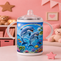 A baby tumbler with clear plastic handles and a clear lid, featuring a colorful underwater print of two dolphins, fish, and a rainbow on a pink-themed room background.