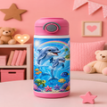 A pink and blue insulated water bottle with a dolphin underwater illustration, set on a pink table in a child's bedroom with teddy bears and a pink wall in the background.