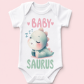 A white short-sleeve baby bodysuit laid flat on a light pink background, featuring a cute cartoon dinosaur graphic with the words 'BABY SAURUS' in pink and mint green text.